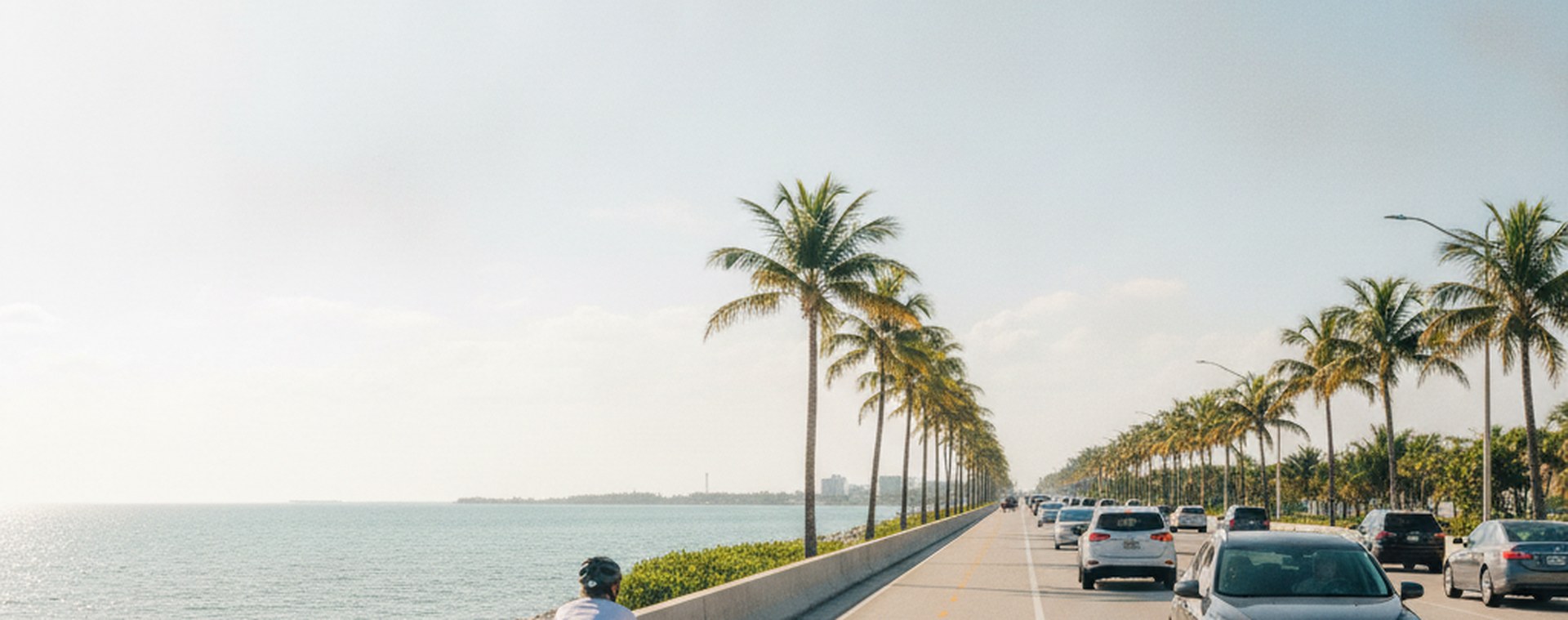 A cyclist riding in a marked bicycle lane on a sunlit Miami road, seen from behind at a distance