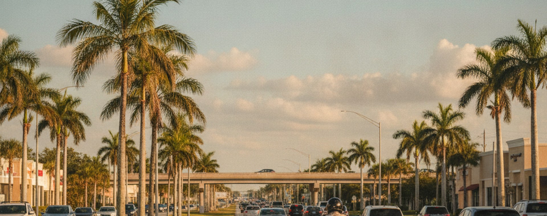 A motorcycle riding in ordinary traffic on a wide Miami road in late-afternoon sun, seen from behind at a distance